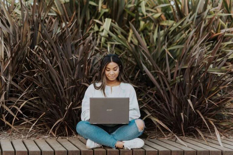 A young female graduate sitting cross-legged outdoors on a wooden deck, working on her laptop with tall ornamental grass in the background, representing a non-EU student searching for employment on a Czech Job Seeker Visa.