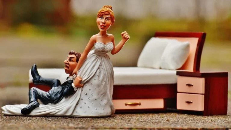 A humorous wedding cake topper figurine featuring a smiling bride in a white dress dragging an anxious groom by his suit collar away from a miniature bed, set against a soft blurred background.