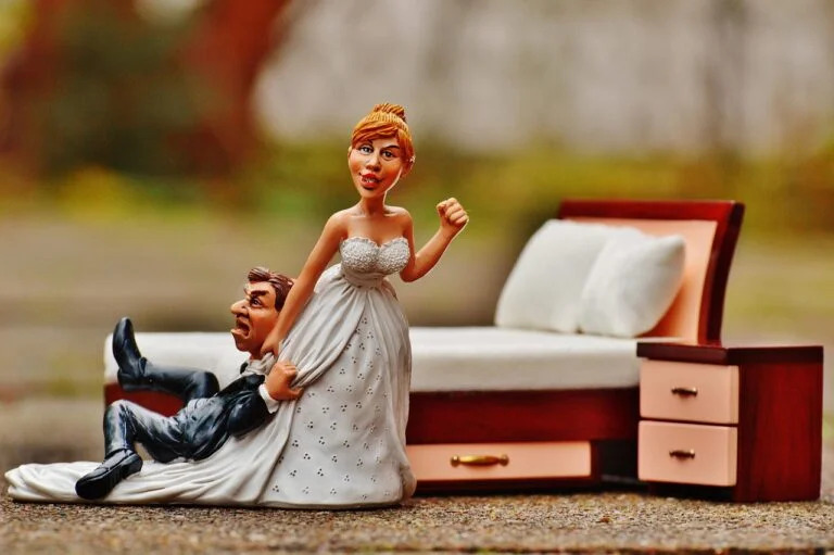 A humorous wedding cake topper figurine featuring a smiling bride in a white dress dragging an anxious groom by his suit collar away from a miniature bed, set against a soft blurred background.
