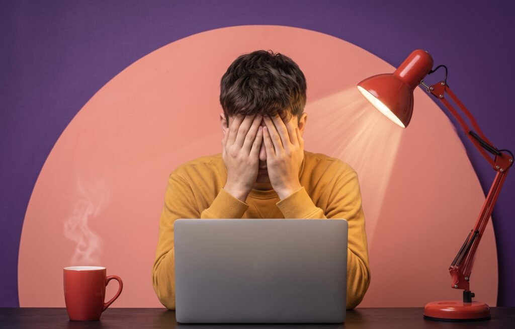 A photo of a young man in a yellow sweater, sitting at a desk and covering his face with his hands in frustration and stress. An open laptop, a red steaming coffee mug, and a red desk lamp are on the desk against a purple and peach geometric wall, symbolizing severe work-related stress and burnout.